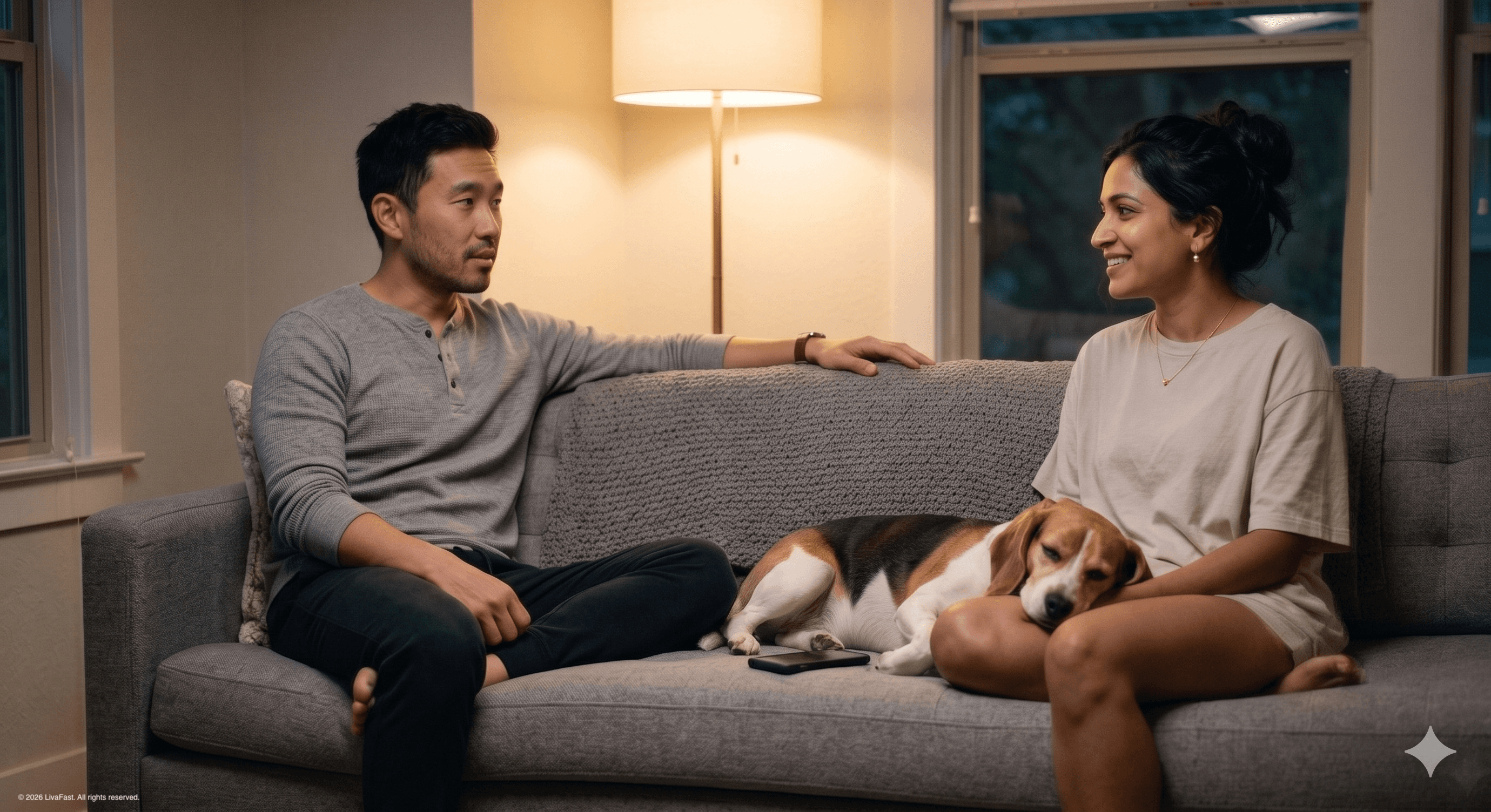 Mark and Priya on a grey sofa in their Austin home in warm evening lamp light, Biscuit the beagle stretched between them, Mark mid-sentence telling her his body is working again.